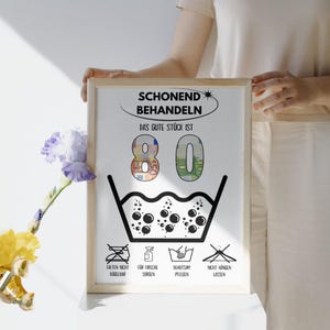 Op de afbeelding: Een witte geframed poster met zwarte tekst die "Schonend Behandeln Das Gute St&uuml;ck Ist 80" luidt. De poster heeft een zwart-witte illustratie van een wasmachine symbool met bubbels en een rij pictogrammen eronder.