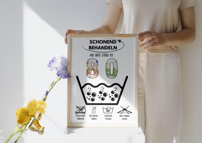 Op de afbeelding: Een witte geframed poster met zwarte tekst die "Schonend Behandeln Das Gute St&uuml;ck Ist 80" luidt. De poster heeft een zwart-witte illustratie van een wasmachine symbool met bubbels en een rij pictogrammen eronder.