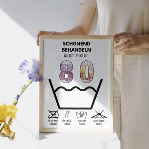 Op de afbeelding: Een wit ingelijst print met de tekst "Schonend Behandeln Das Gute St&uuml;ck Ist 80" en een zwart-witte illustratie van een wasbak met water en een streep erdoor. Onder de bak bevinden zich vier pictogrammen: een strijkijzer met een streep erdoor, een fles met een waterdruppel, een emmer met een hand erin en een hanger met een streep erdoor.
