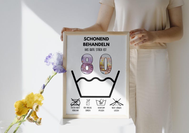 Op de afbeelding: Een wit ingelijst print met de tekst "Schonend Behandeln Das Gute St&uuml;ck Ist 80" en een zwart-witte illustratie van een wasbak met water en een streep erdoor. Onder de bak bevinden zich vier pictogrammen: een strijkijzer met een streep erdoor, een fles met een waterdruppel, een emmer met een hand erin en een hanger met een streep erdoor.