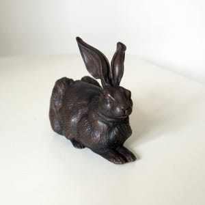 May include: A bronze-colored rabbit figurine in a relaxed pose. The sculpture features detailed fur texture and long, upright ears. The rabbit is resting on its front paws, creating a lifelike appearance. This decorative item is suitable for home decor.