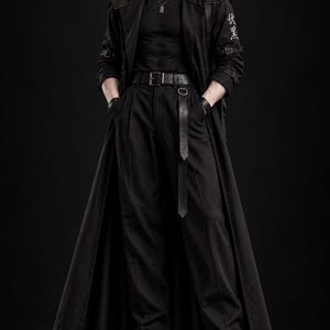 May include: A male anime-style figurine wearing a black long coat, black shirt, and black trousers. The figure features a silver dog tag necklace, a black belt with a long strap, and black boots. A rectangular case is strapped to the back.