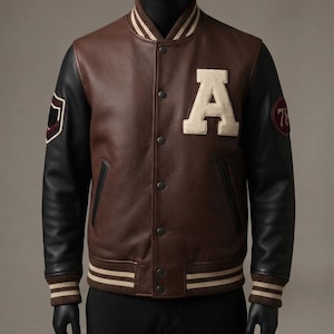 Op de afbeelding: Een bruin leren varsity-jack met zwarte leren mouwen, voorzien van een grote crèmekleurige letter A op de borst. Het jack heeft gestreepte geribbelde boorden bij de mouwen en zoom, drukknoopsluitingen en ronde patches op beide mouwen.