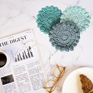 May include: Three crocheted coasters in shades of blue and green. The coasters are round and have a textured surface. They are arranged on a white marble surface.