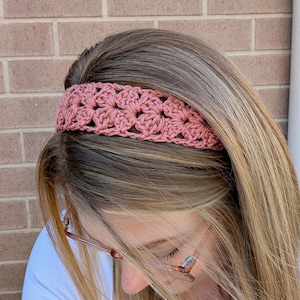 May include: A dusky pink crochet headband featuring a floral pattern. The headband, made from soft yarn, is worn across the forehead. This hair accessory is a stylish addition to any outfit, measuring approximately 5 cm in width.
