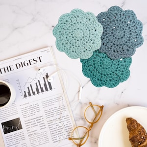 May include: Three crocheted coasters in shades of blue and green. The coasters are round and have a scalloped edge. They are arranged on a white marble surface.