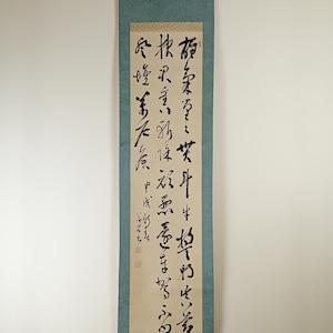 Può includere: Un dipinto su rotolo giapponese tradizionale con calligrafia di inchiostro nero su sfondo beige. Il rotolo è montato su un supporto in tessuto blu.