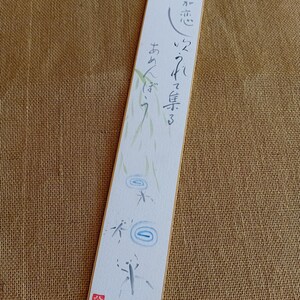 Puede incluir: Un marcapáginas rectangular con caligrafía japonesa e ilustraciones en acuarela. El marcapáginas blanco tiene un borde dorado y presenta texto, bambú y diseños de gotas de agua. Un sello rojo está en la esquina inferior izquierda.