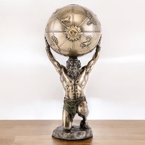 May include: A bronze-effect statue of Atlas kneeling and supporting a globe. The globe is decorated with zodiac symbols and a sun. The statue is on a stone base, placed on a wooden surface.
