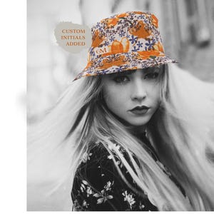 May include: A floral bucket hat with orange and blue accents. The hat features a pattern of flowers and leaves. The text "CUSTOM INITIALS ADDED" is visible. The initials "GM" are also visible on the hat.