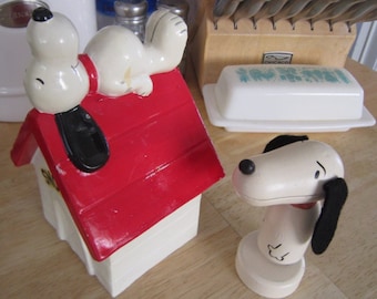 May include: A red and white ceramic dog house with a white ceramic dog laying on the roof. A second white ceramic dog with black ears is standing on a white base.