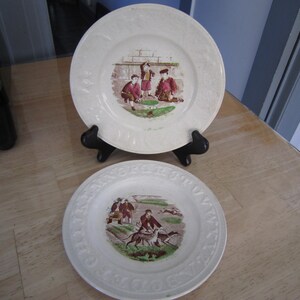 May include: Two white ceramic plates with a decorative border. The plates feature a central image of children playing games. One plate shows children playing with a ball, while the other shows children playing with dogs.
