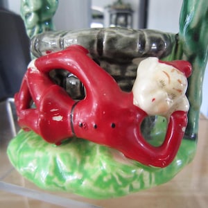 May include: A ceramic figurine of a sleeping elf in red and green clothing, lying on a green base with a stone well behind it.