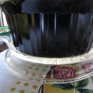Puede incluir: Un plato para hornear de vidrio negro con una tapa de vidrio transparente y una base de vidrio transparente. El plato est&aacute; sobre un mantel blanco con un patr&oacute;n floral.