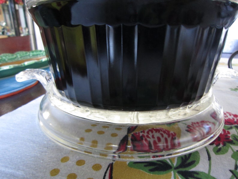 Puede incluir: Un plato para hornear de vidrio negro con una tapa de vidrio transparente y una base de vidrio transparente. El plato est&aacute; sobre un mantel blanco con un patr&oacute;n floral.