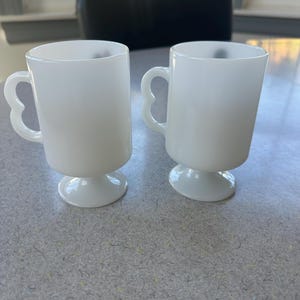 May include: Two white milk glass mugs with a smooth, opaque finish. Each mug features a cylindrical body, a pedestal base, and a distinctive heart-shaped handle. The mugs are identical in design, standing upright with a clean, simple aesthetic.