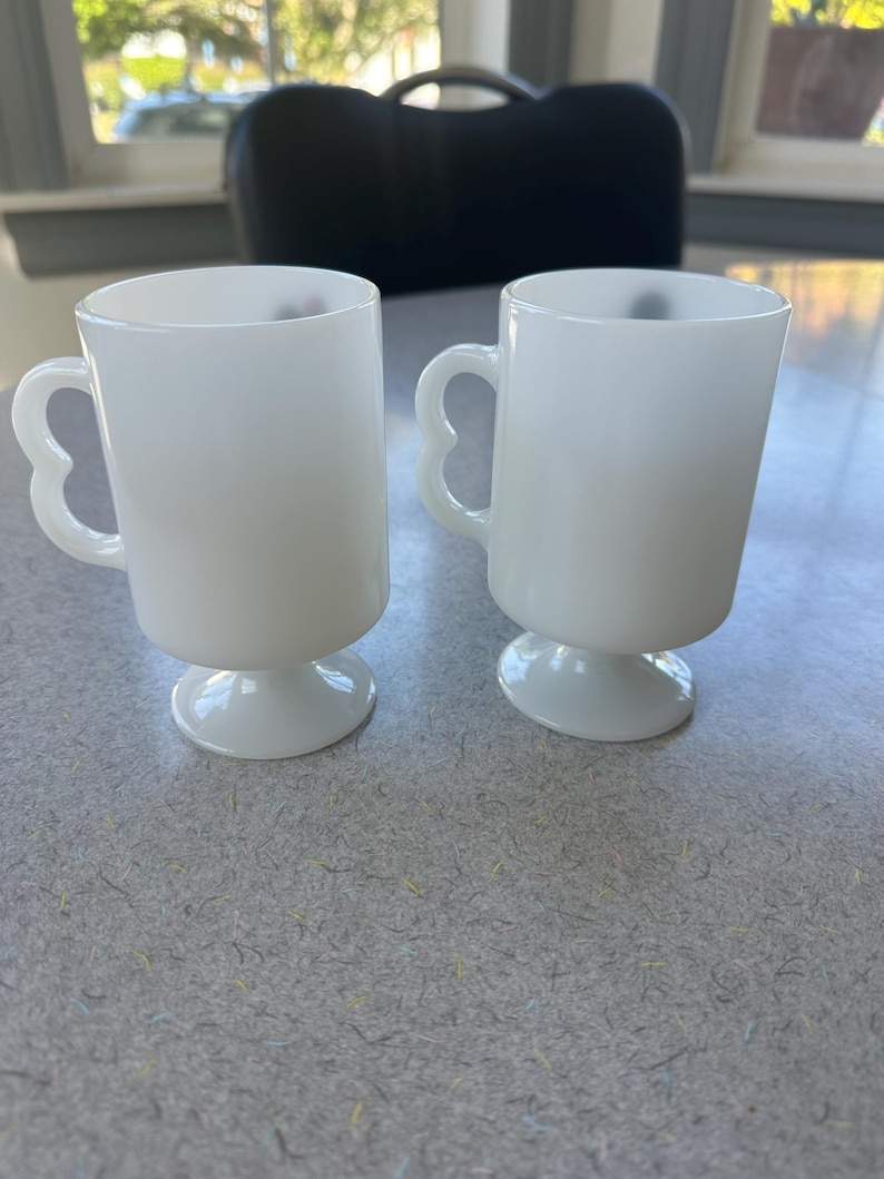 May include: Two white milk glass mugs with a smooth, opaque finish. Each mug features a cylindrical body, a pedestal base, and a distinctive heart-shaped handle. The mugs are identical in design, standing upright with a clean, simple aesthetic.