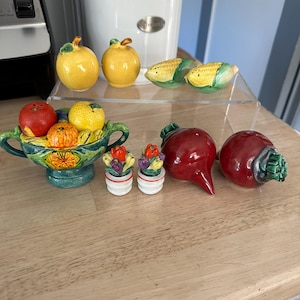 May include: A collection of ceramic salt and pepper shakers in the shapes of fruits and vegetables. Includes yellow lemons, corn on the cob, red beets, and a decorative bowl filled with fruit. The shakers are brightly colored and detailed.