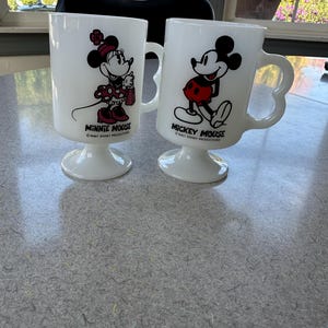 May include: Two white milk glass pedestal mugs featuring Disney characters. One mug displays Minnie Mouse with the text MINNIE MOUSE WALT DISNEY PRODUCTIONS. The other mug displays Mickey Mouse with the text MICKEY MOUSE WALT DISNEY PRODUCTIONS.