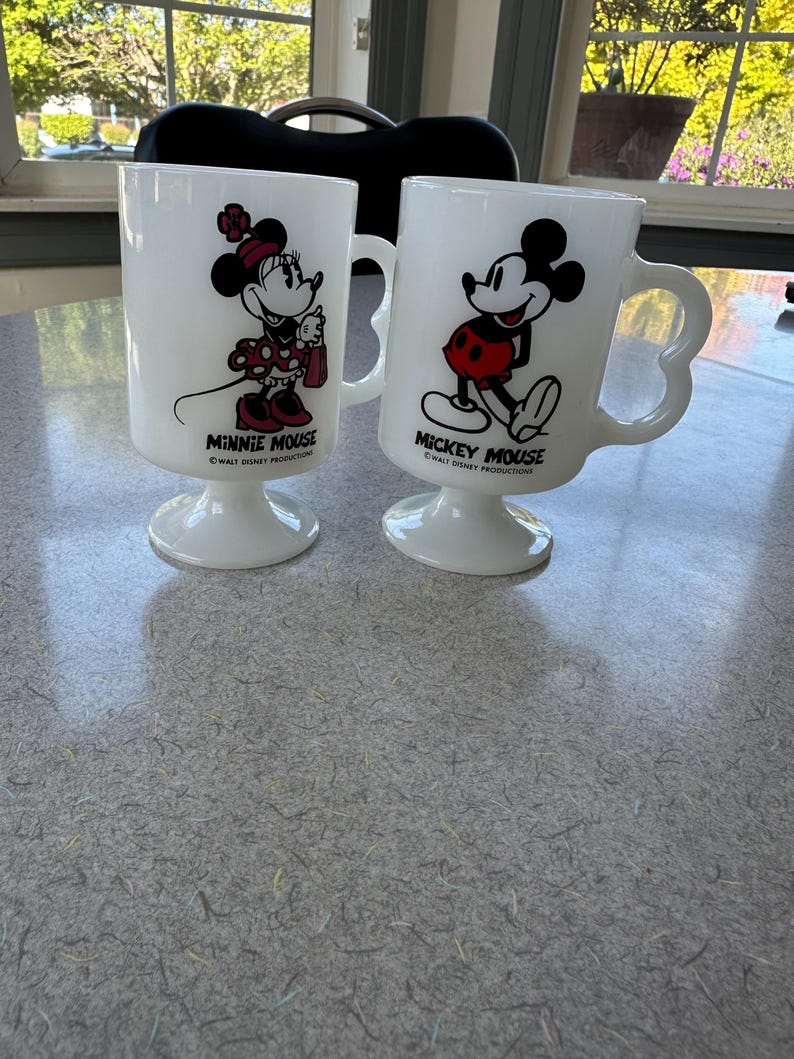 May include: Two white milk glass pedestal mugs featuring Disney characters. One mug displays Minnie Mouse with the text MINNIE MOUSE WALT DISNEY PRODUCTIONS. The other mug displays Mickey Mouse with the text MICKEY MOUSE WALT DISNEY PRODUCTIONS.