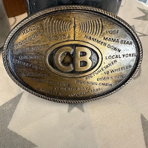 May include: Oval-shaped, antique-style belt buckle with a detailed design. The center features the letters "CB" in a bold font. The buckle is inscribed with phrases like "Handle," "Mama Bear," and "18 Wheeler."