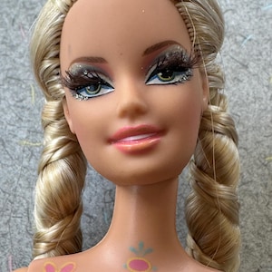 May include: A close-up of a doll's face with blonde hair styled in two braids. The doll has detailed makeup, including long eyelashes, glitter, and pink lipstick. The doll's skin has painted designs on the chest and shoulder.