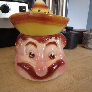 May include: A ceramic cookie jar shaped like a man's head wearing a yellow sombrero. The man has a brown mustache and a wide smile.
