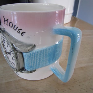 Puede incluir: Una taza de cer&aacute;mica blanca con un dise&ntilde;o rosa y azul. La taza presenta un reloj 3D con una banda azul y la palabra "Mouse" escrita en el lateral.