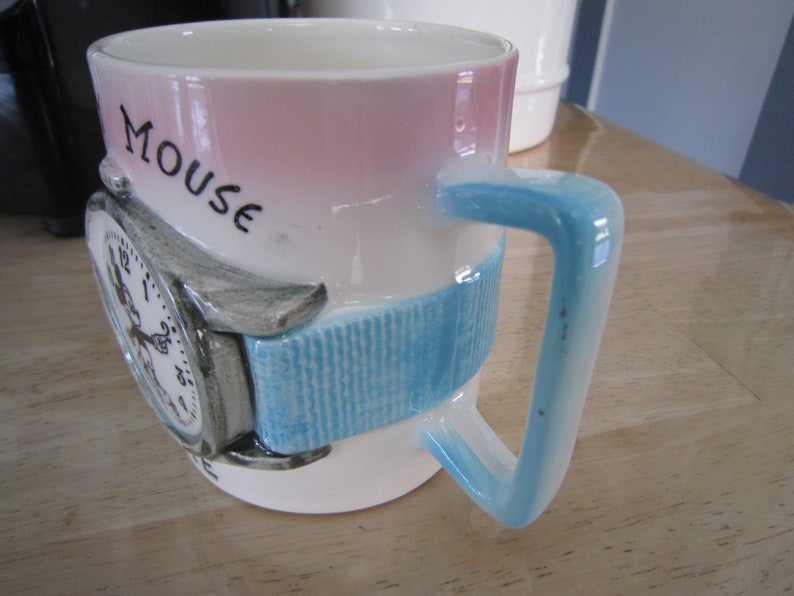 Puede incluir: Una taza de cer&aacute;mica blanca con un dise&ntilde;o rosa y azul. La taza presenta un reloj 3D con una banda azul y la palabra "Mouse" escrita en el lateral.
