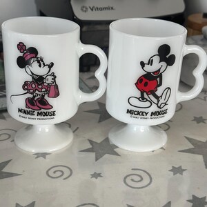 May include: Two white milk glass pedestal mugs featuring heart-shaped handles. One mug displays Minnie Mouse with pink accents and the text Minnie Mouse Walt Disney Productions. The other mug shows Mickey Mouse in red shorts with the text Mickey Mouse Walt Disney Productions.