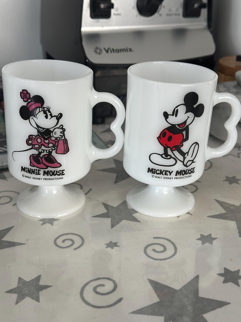 May include: Two white milk glass pedestal mugs featuring heart-shaped handles. One mug displays Minnie Mouse with pink accents and the text Minnie Mouse Walt Disney Productions. The other mug shows Mickey Mouse in red shorts with the text Mickey Mouse Walt Disney Productions.