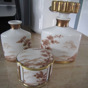 May include: A set of three white porcelain bottles with gold accents and brown painted landscapes. The bottles have a Japanese-inspired design.