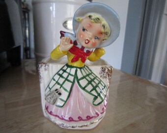 May include: A vintage ceramic planter shaped like a girl wearing a blue and white hat, a yellow and red plaid dress, and a pink skirt. The planter has a gold trim and is decorated with a floral pattern.