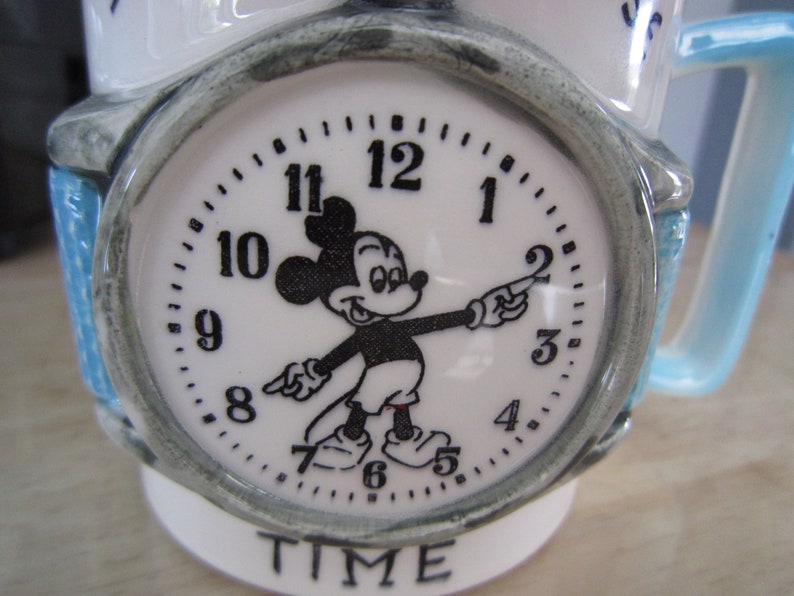 Puede incluir: Taza de cer&aacute;mica blanca con asa azul. La taza presenta una esfera de reloj de Mickey Mouse con las palabras "TIME" impresas debajo.