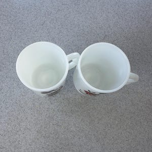 May include: Two white milk glass mugs viewed from above, each featuring a side handle and a partial graphic design on the exterior. The mugs have a smooth, opaque finish and a cylindrical shape, showing the hollow interior of each vessel clearly.