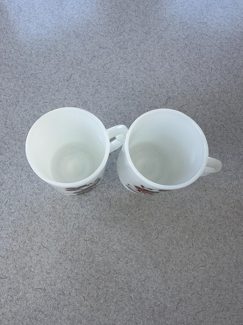 May include: Two white milk glass mugs viewed from above, each featuring a side handle and a partial graphic design on the exterior. The mugs have a smooth, opaque finish and a cylindrical shape, showing the hollow interior of each vessel clearly.