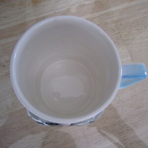 Puede incluir: Una taza de cer&aacute;mica blanca con un asa azul. La taza est&aacute; vac&iacute;a y tiene un interior blanco liso.