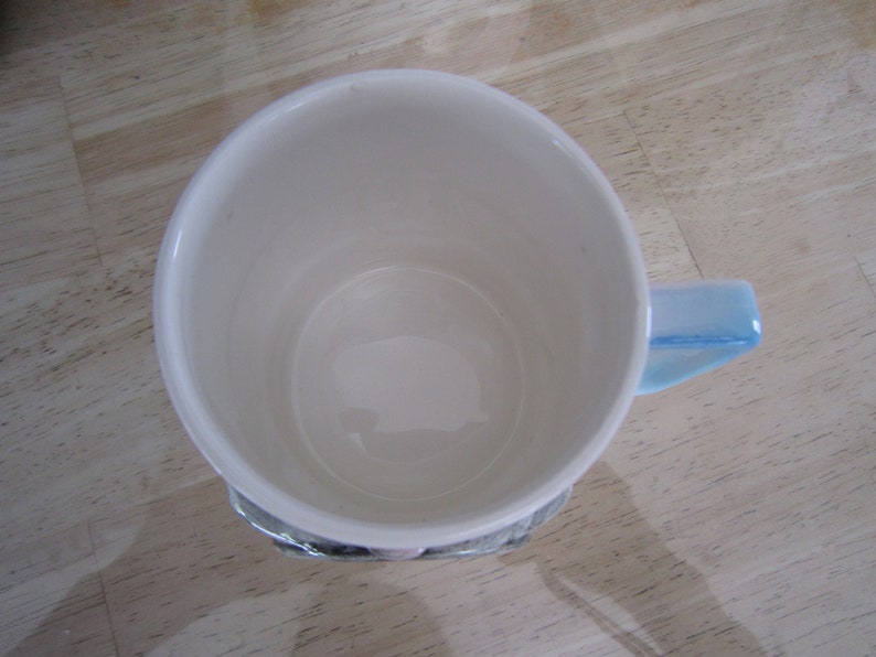 Puede incluir: Una taza de cer&aacute;mica blanca con un asa azul. La taza est&aacute; vac&iacute;a y tiene un interior blanco liso.