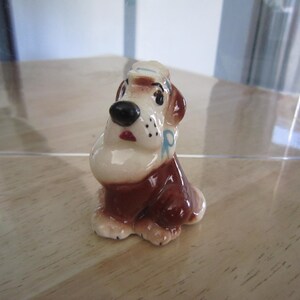May include: A ceramic figurine of a brown and white dog wearing a blue and white sailor hat. The dog has a blue ribbon around its neck and is sitting with its head tilted to the side.