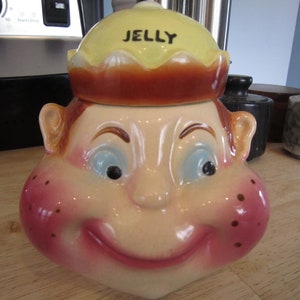 May include: A ceramic jelly jar shaped like a smiling face with rosy cheeks and brown hair. The lid is yellow with the word "JELLY" written in black.