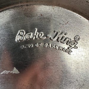 May include: Close-up of a well-worn, circular metal baking pan. The embossed text "Bake-King" is visible, along with "3310 OF BAKEWARE". The pan has a textured surface with signs of use, suggesting it's a vintage kitchen item.