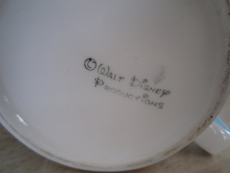 Puede incluir: Primer plano de la parte inferior de una taza de cer&aacute;mica blanca con texto negro que dice "&copy; WALT DISNEY PRODUCTIONS".