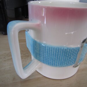 Puede incluir: Taza de cer&aacute;mica blanca con un dise&ntilde;o degradado azul y rosa. La taza tiene una banda azul con un patr&oacute;n texturizado alrededor del medio y un detalle de hebilla plateada.