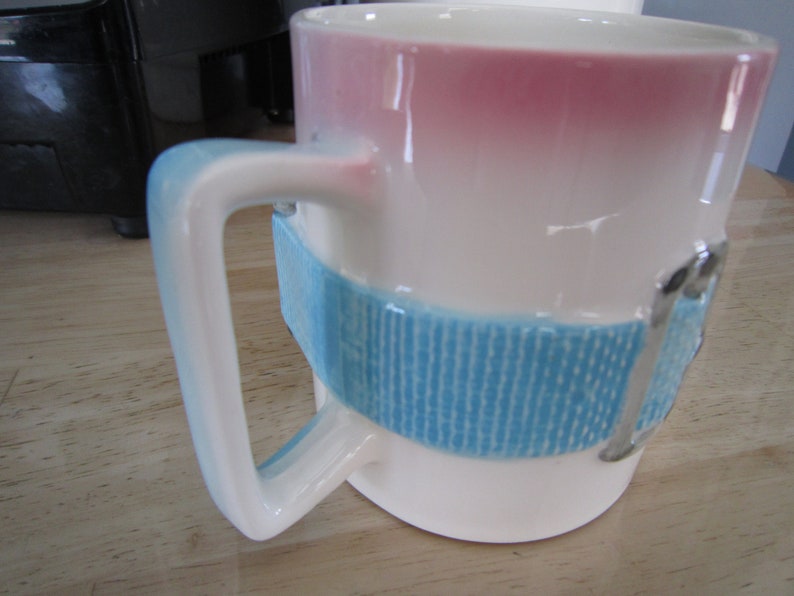 Puede incluir: Taza de cer&aacute;mica blanca con un dise&ntilde;o degradado azul y rosa. La taza tiene una banda azul con un patr&oacute;n texturizado alrededor del medio y un detalle de hebilla plateada.