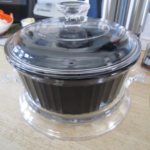 Puede incluir: Un plato de vidrio transparente con un interior negro y una tapa de vidrio. El plato tiene dos asas a los lados y se asienta sobre una base de vidrio transparente.