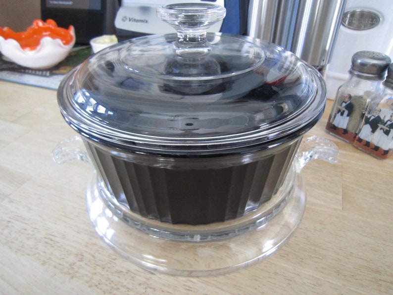 Puede incluir: Un plato de vidrio transparente con un interior negro y una tapa de vidrio. El plato tiene dos asas a los lados y se asienta sobre una base de vidrio transparente.
