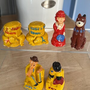 May include: A collection of vintage ceramic figurines. The set includes yellow salt and pepper shakers with dog figures, a red-dressed woman, a brown dog with a blue collar, and two yellow figures with hats. The figurines are displayed on a clear shelf.