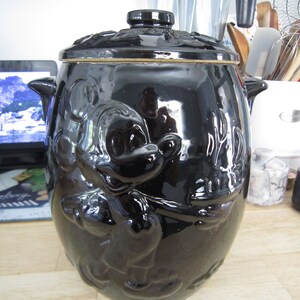 May include: Black ceramic cookie jar with a Mickey Mouse design. The jar has a lid and is decorated with a brown rim.