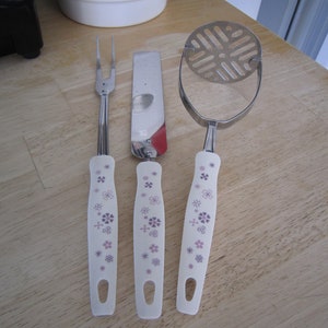 May include: Three kitchen utensils with white handles featuring a floral pattern in shades of purple and pink. The utensils are a fork, a spatula, and a potato masher.