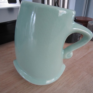 May include: A light green ceramic mug with a unique, curved handle. The mug is sitting on a wooden surface.